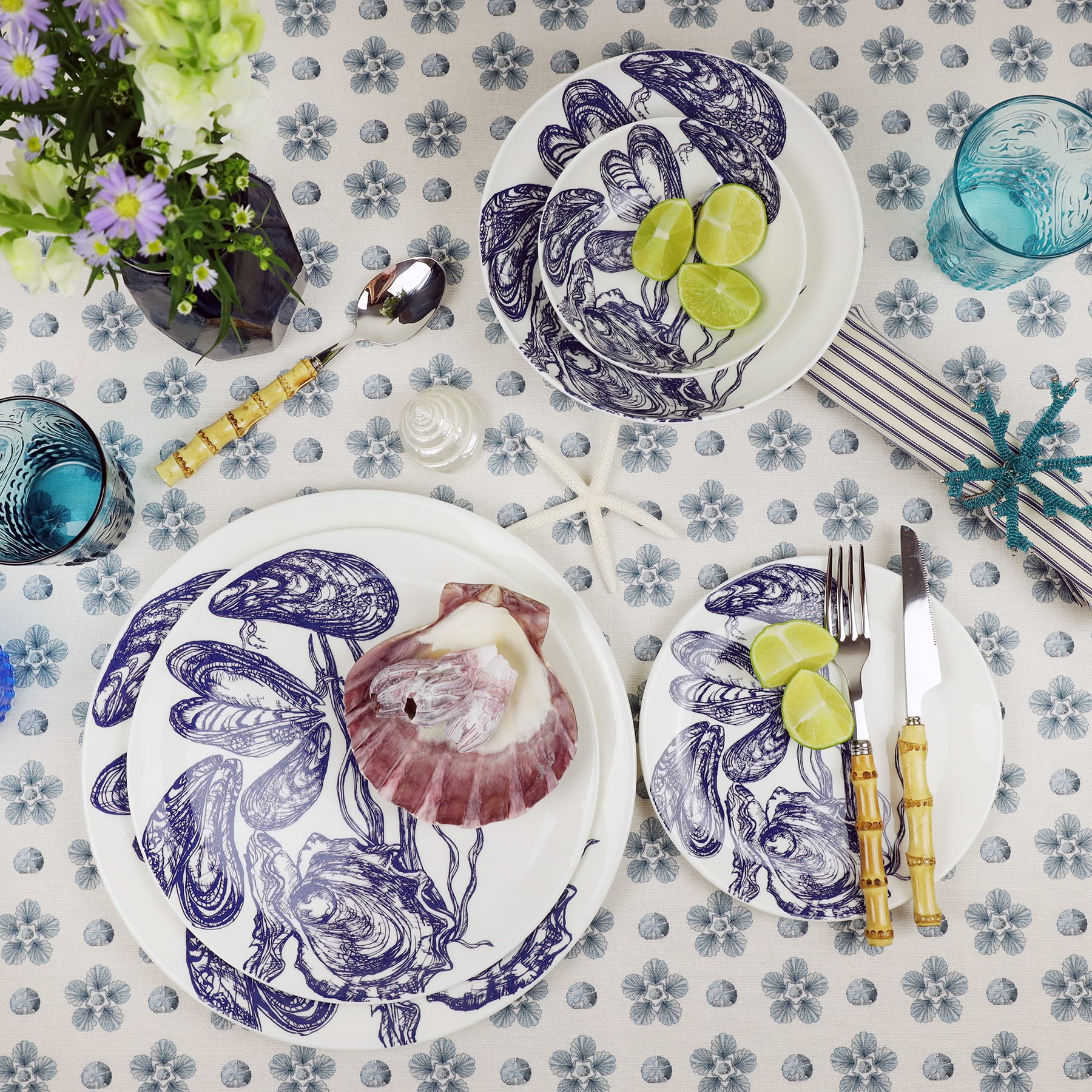 Pasta bowl in Bone China in our Classic range in Navy and white in the Mussel design, and a smaller bowl placed inside with a couple of limes.All placed on mini star fabric,next to the bowl is a mussel dinner plate with a scallop shell on top.A side plate has bamboo cutlery and a couple of limes on.On the table is a vase with flowers 