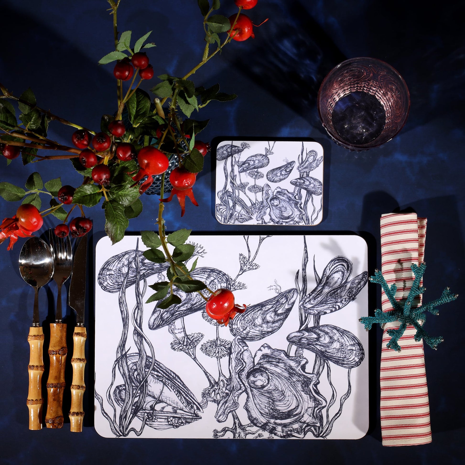 Mussel and Oyster Design in Navy on a white Coaster with a matching Placemat in a table setting on a dark table cloth.On the table is bamboo cutlery, stripe napkin and a glass with rosehips.