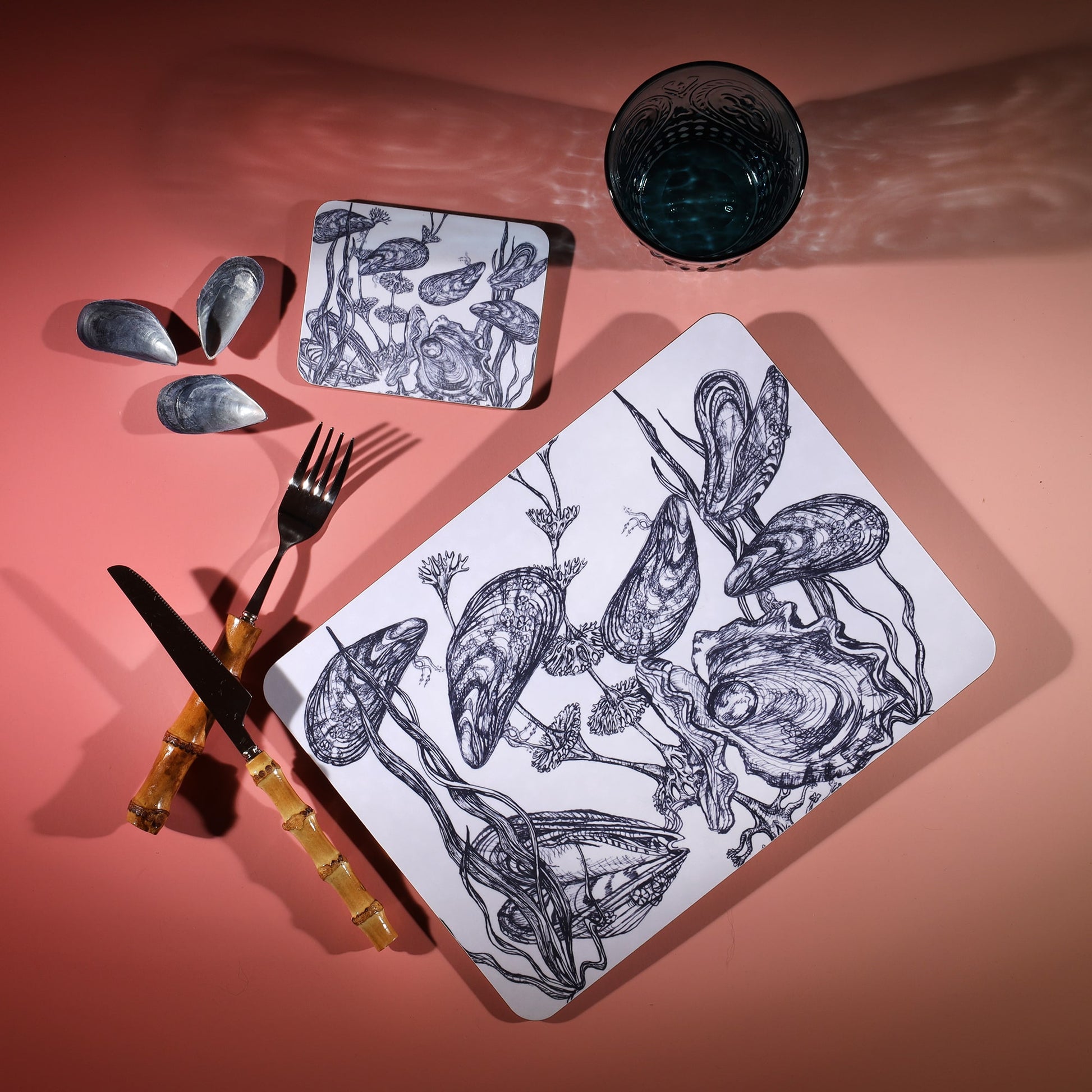 Mussel  Design in Navy on a white Coaster with a matching Placemat.On the table is a bamboo cutlery set and a blue coloured glass