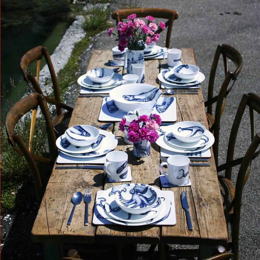 Table set by the sea with six place settings of our Bone China tableware on one of our tablecloths.On the table are two jugs with sweet peas and coloured glasses decorate the table.You can see the sea in the background