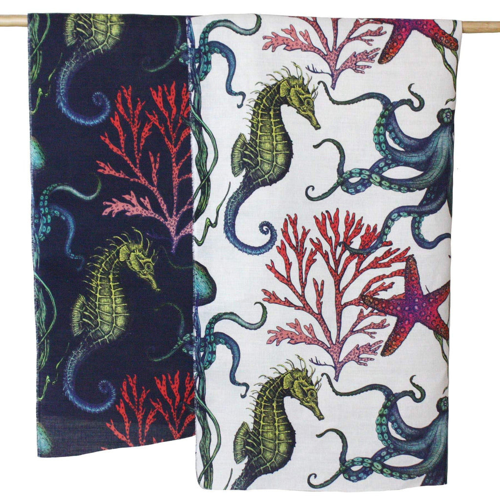 Two panels of Cream Cornwall's Rainbow Reef fabric hanging side by side. The left fabric features a deep navy background, while the right fabric has a white background. Both showcase detailed illustrations of green seahorses, purple and red coral, vibrant starfish, and teal-blue octopuses, creating a vivid underwater scene.