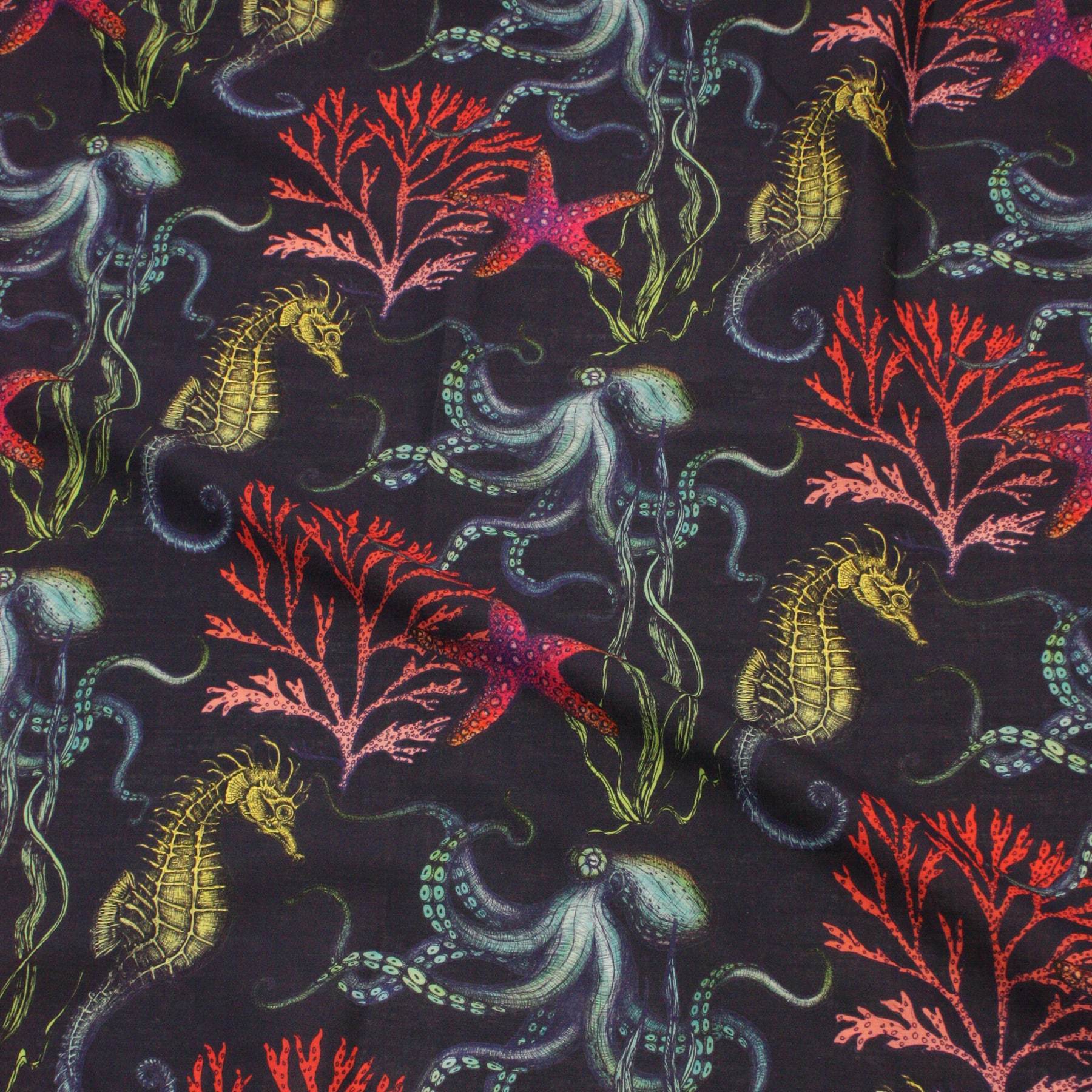Flat lay of Cream Cornwall’s Rainbow Reef fabric in navy, showcasing an intricate underwater design with vibrant illustrations of seahorses, octopuses, starfish, and coral in shades of red, purple, green, and teal against a rich dark background.