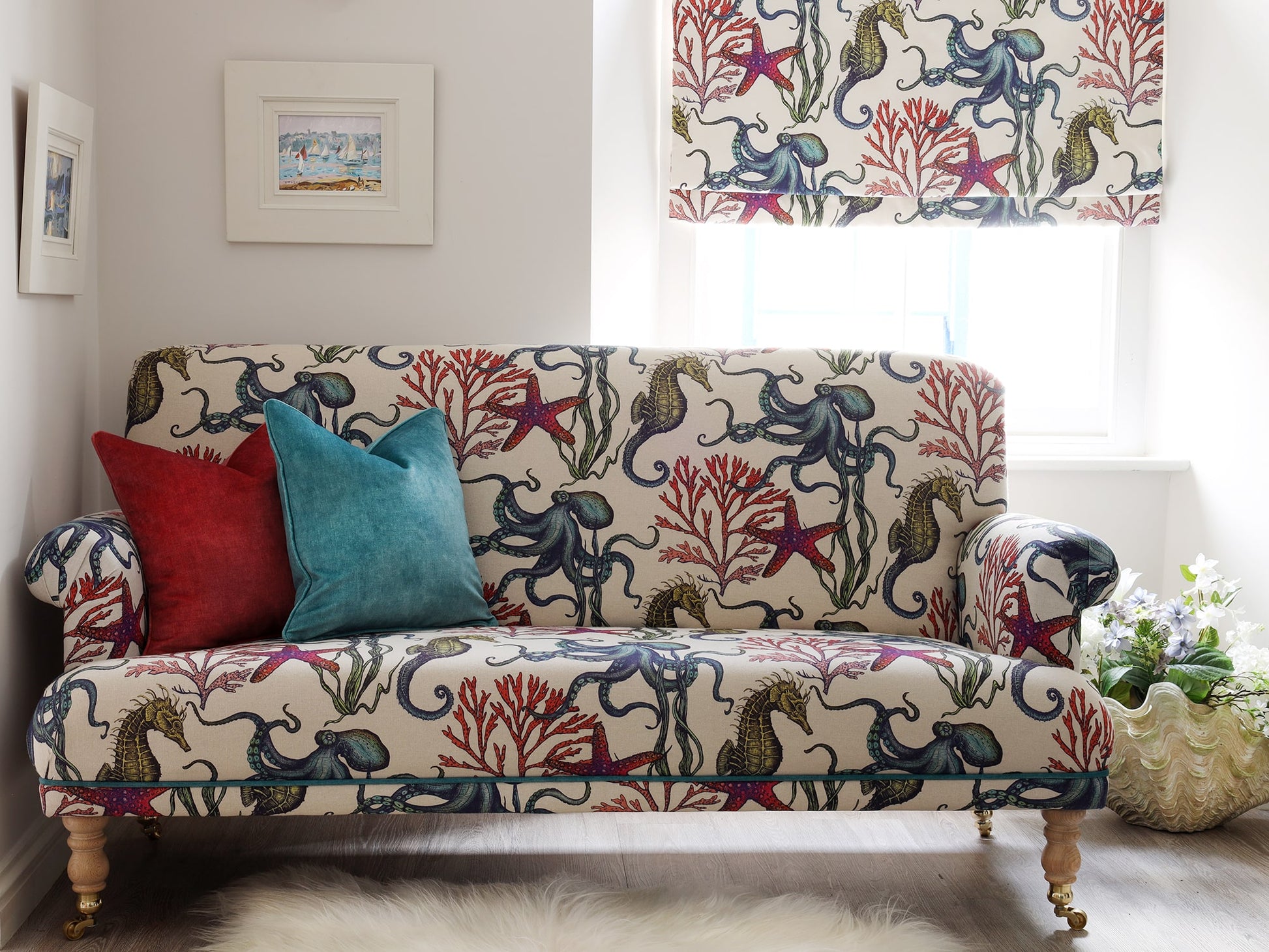 A bright and coastal-inspired living room featuring a cream-colored sofa and matching Roman blind, both upholstered in Cream Cornwall’s Rainbow Reef fabric. The vibrant marine design includes octopuses, seahorses, starfish, and coral in rich blues, reds, and greens. The sofa is accessorized with plush red and teal velvet cushions, and a large clam shell planter with white flowers sits by the window, creating a fresh, seaside ambiance.