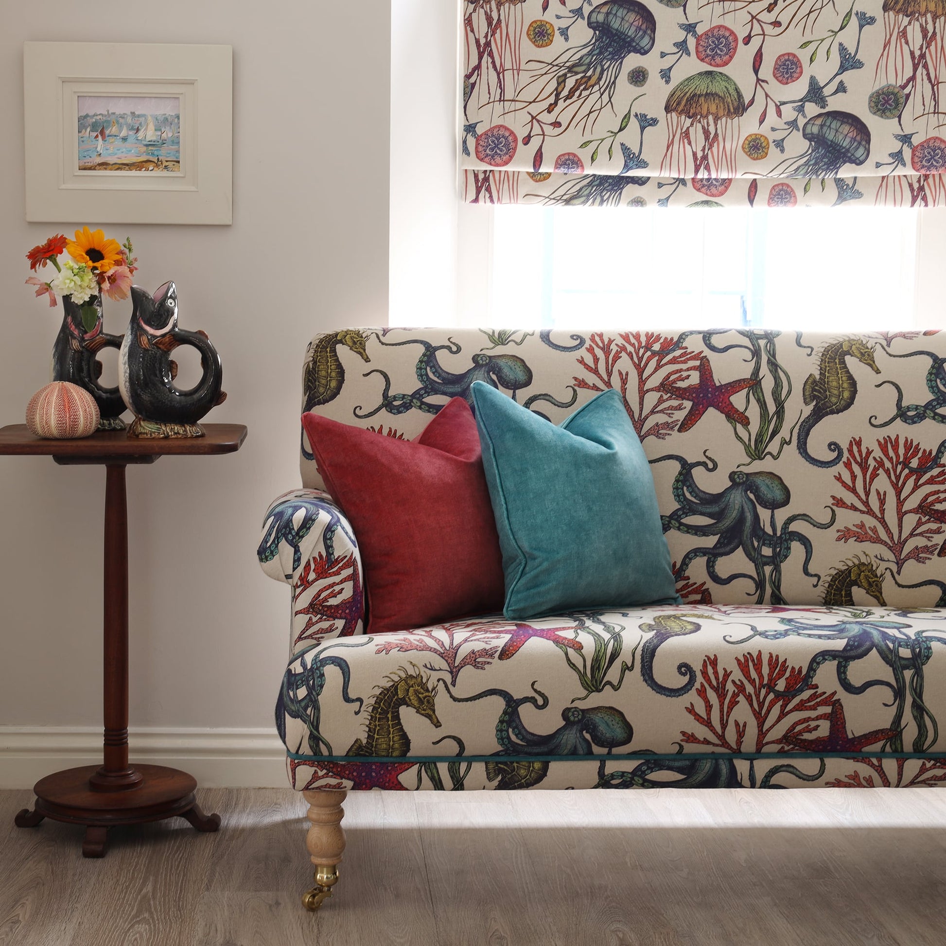 A cheerful coastal-themed interior featuring a sofa upholstered in Cream Cornwall's Rainbow Reef fabric in natural tones. The upholstery is adorned with detailed illustrations of seahorses, octopuses, starfish, and coral in vibrant shades. Two velvet cushions in deep red and teal add a pop of colour. Beside the sofa is a small wooden side table with a whimsical fish-shaped jug holding a bouquet of fresh flowers, next to a Roman blind featuring Cream Cornwall’s jellyfish-themed Canyons Reef design.