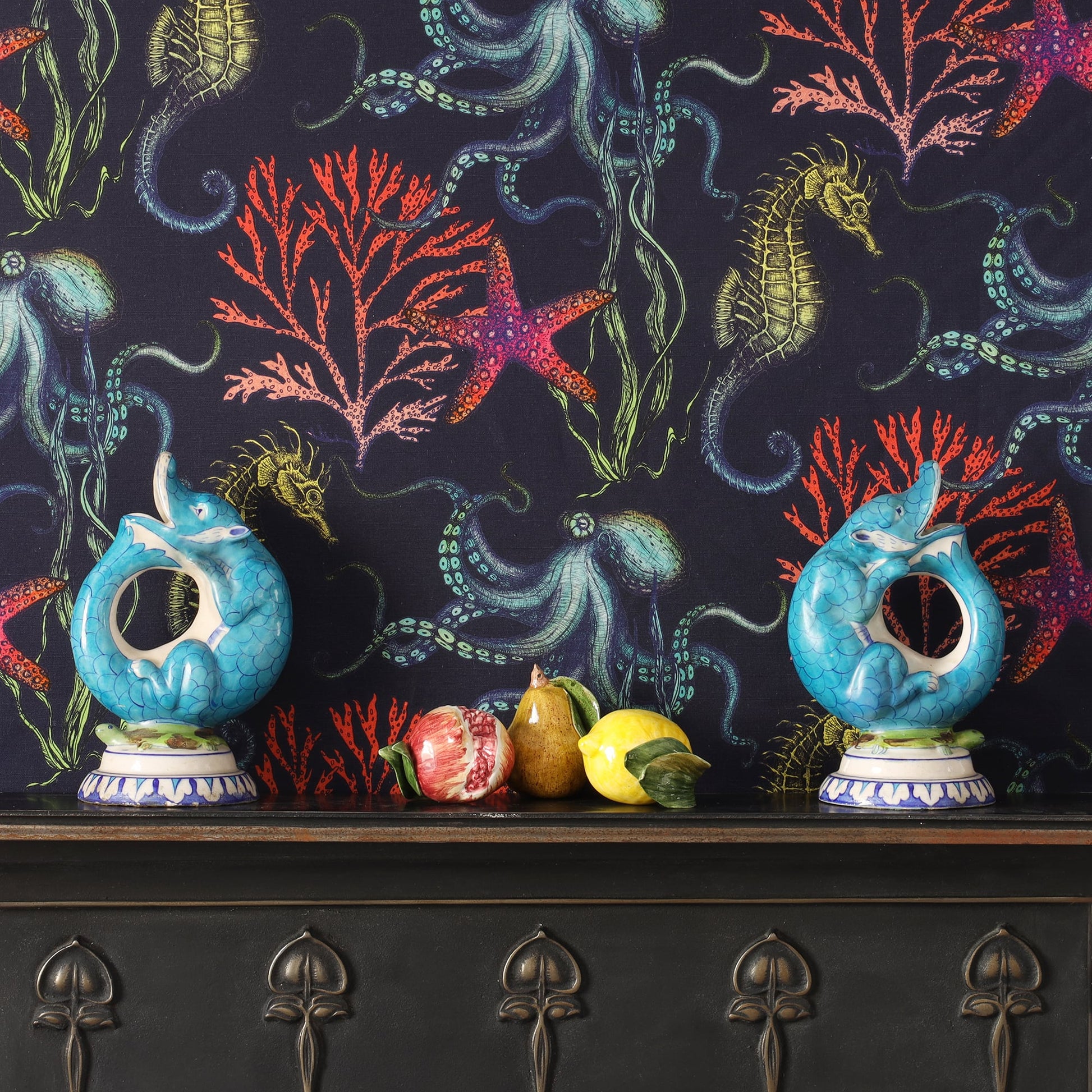 A rich navy Rainbow Reef fabric from Cream Cornwall used as a backdrop, featuring vibrant underwater illustrations of octopuses, seahorses, coral, and starfish in bold shades of turquoise, red, and gold. In front sits a dark wooden sideboard adorned with two turquoise ceramic fish vases and ceramic fruits including a pear, lemon, and pomegranate.