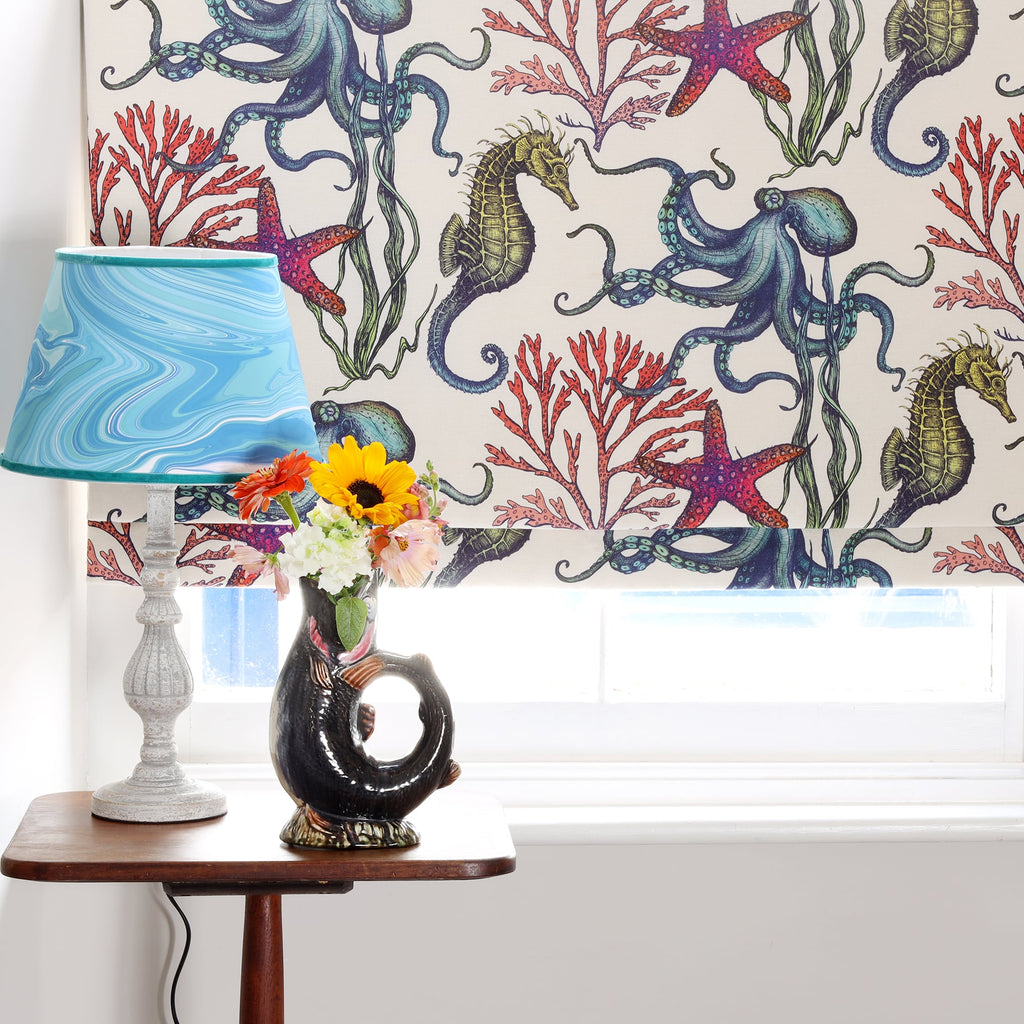 A window dressed with Cream Cornwall's Rainbow Reef fabric blind in off-white, featuring detailed illustrations of octopuses, seahorses, starfish, and red coral. Below the window sits a small wooden table with a ceramic fish jug vase filled with vibrant fresh flowers and a lamp with a swirling blue lampshade, creating a cheerful coastal-inspired interior scene.