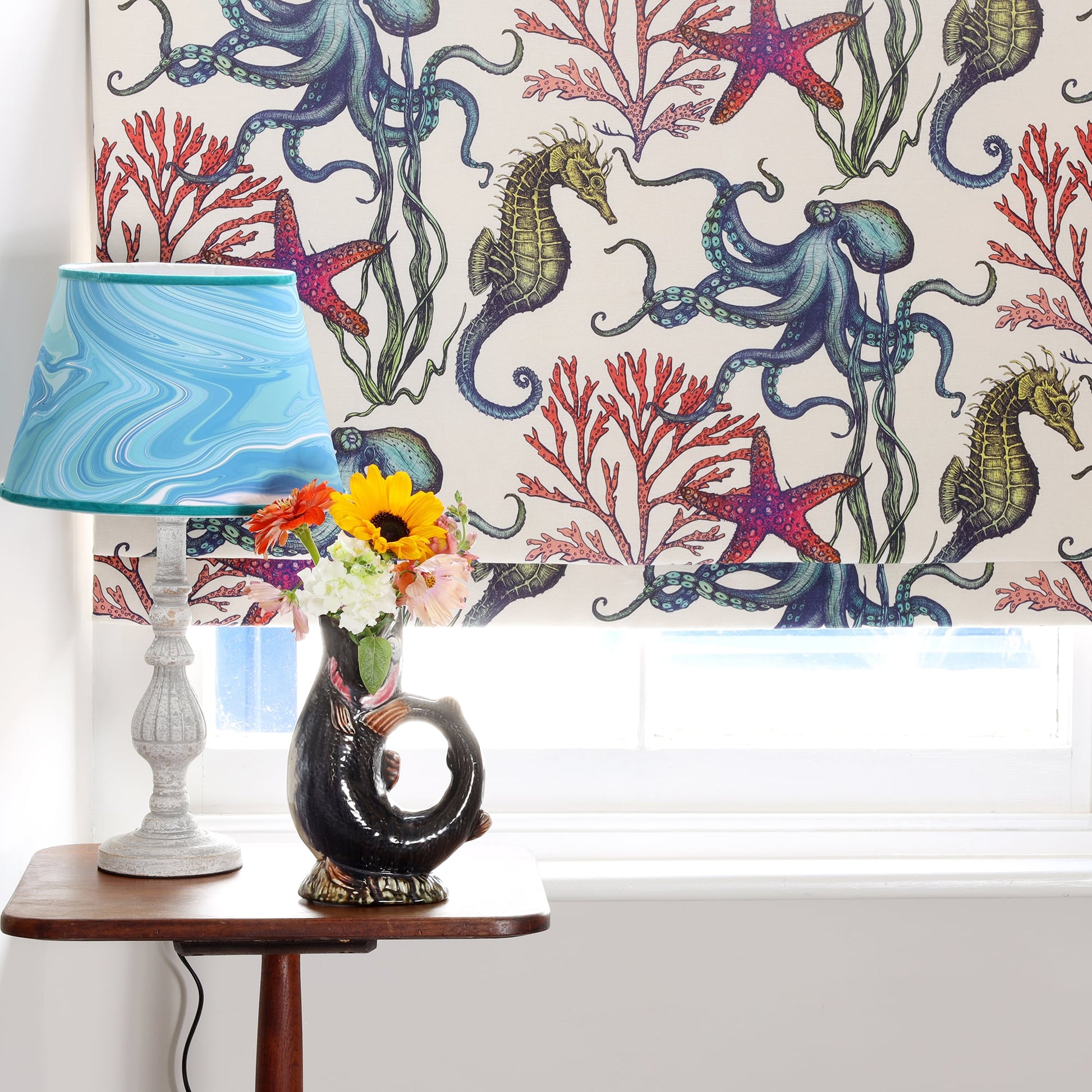 A window dressed with Cream Cornwall's Rainbow Reef fabric blind in off-white, featuring detailed illustrations of octopuses, seahorses, starfish, and red coral. Below the window sits a small wooden table with a ceramic fish jug vase filled with vibrant fresh flowers and a lamp with a swirling blue lampshade, creating a cheerful coastal-inspired interior scene.