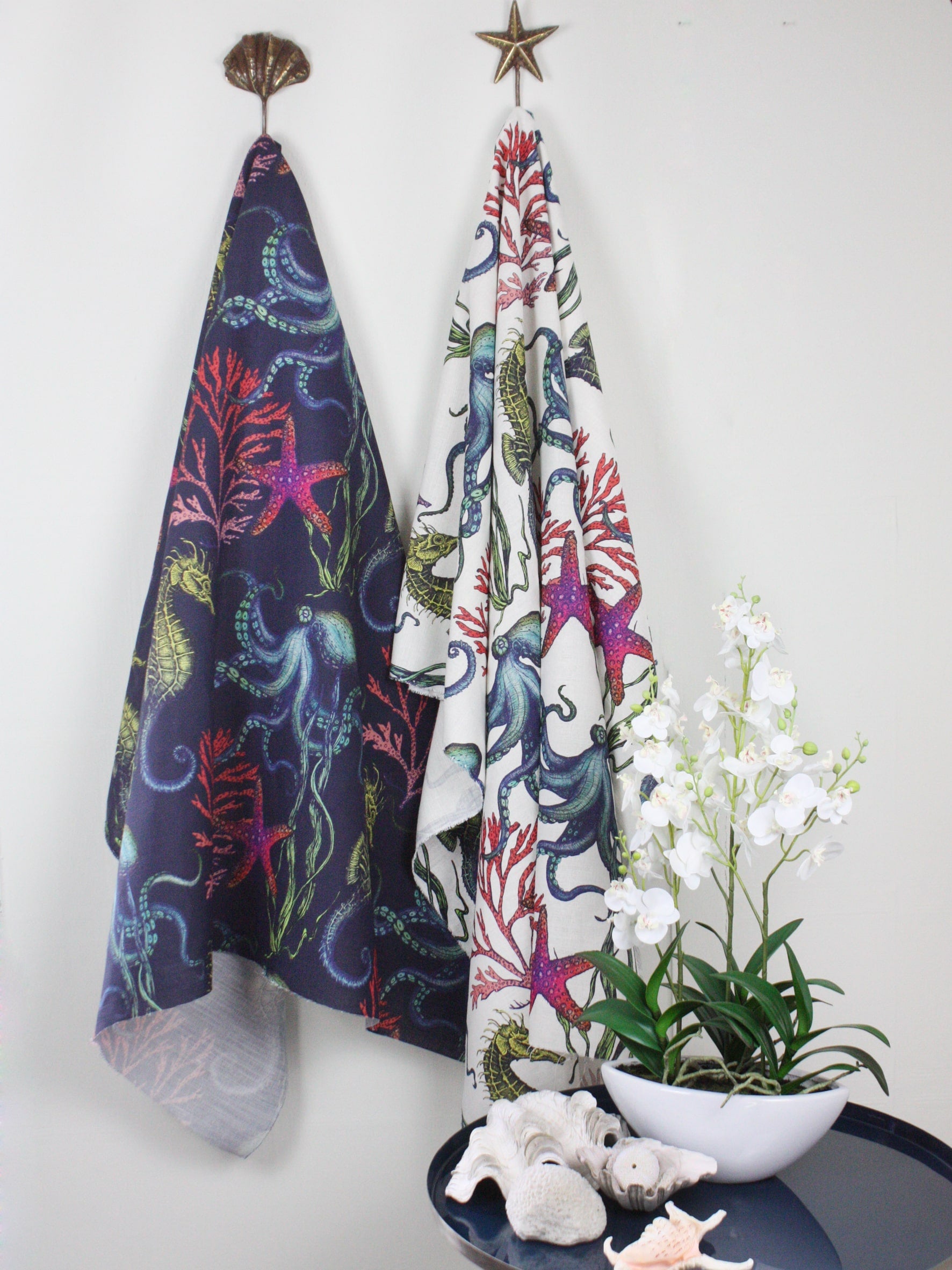 Two lengths of Cream Cornwall's Rainbow Reef fabric hanging on decorative wall hooks—one with a deep navy background and the other with a crisp white background. Both fabrics feature vibrant illustrations of octopuses, seahorses, starfish, and coral. Below, a navy side table is styled with white seashells and a white orchid plant in a bowl.