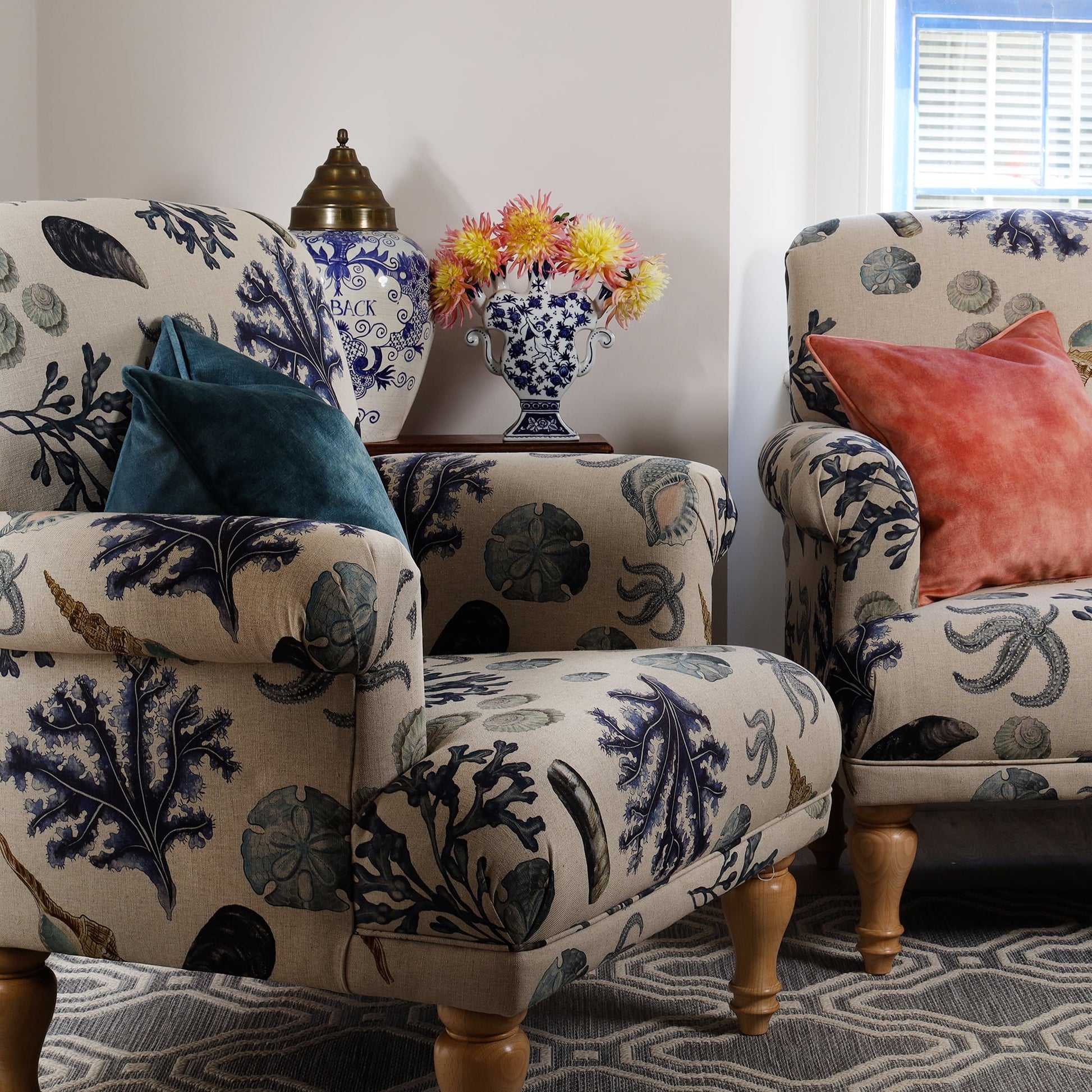 A cosy sitting area with a matching armchair and loveseat upholstered in Cream Cornwall's Rockpool fabric in natural linen. The design features seaweed, shells, and marine life in soft coastal tones. A teal cushion sits on the armchair, with a coral cushion on the loveseat. A side table holds a blue and white vase with dahlias, adding a coastal charm.