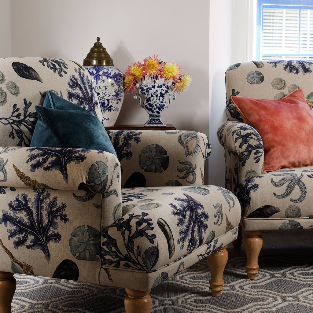A cosy sitting room featuring a matching armchair and loveseat upholstered in Cream Cornwall's Rockpool fabric in a natural linen shade. The fabric is adorned with detailed illustrations of seaweed, seashells, and marine life in muted coastal blues and soft greys. A teal velvet cushion rests on the armchair, while the loveseat is accented with a coral velvet cushion. Behind the armchair is a dark wooden side table topped with a blue and white ceramic vase filled with pink and yellow dahlias.