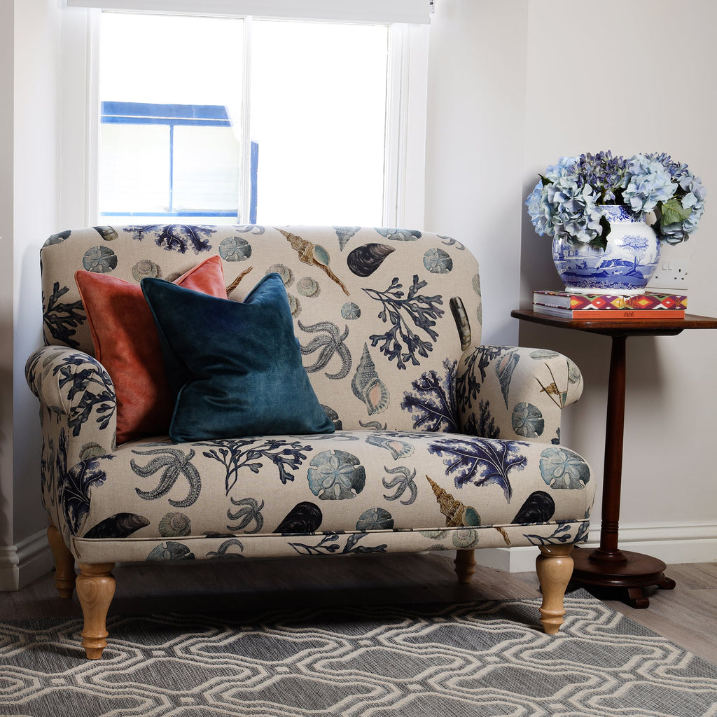 A small loveseat upholstered in Cream Cornwall's Rockpool natural fabric, featuring sea-themed illustrations in soft blue and neutral tones. Two velvet cushions in teal and coral add a pop of colour. A wooden side table holds a blue and white vase with hydrangeas.
