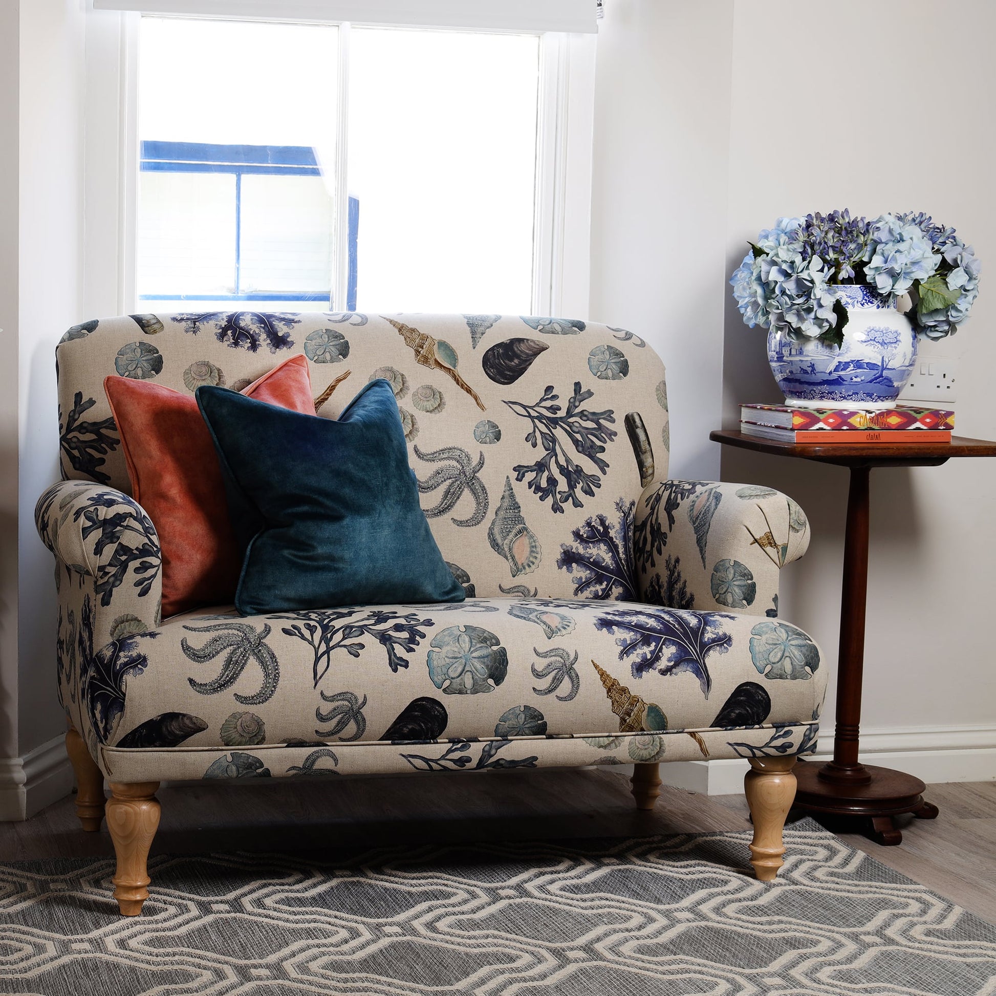 A small loveseat upholstered in Cream Cornwall's Rockpool natural fabric, featuring sea-themed illustrations in soft blue and neutral tones. Two velvet cushions in teal and coral add a pop of colour. A wooden side table holds a blue and white vase with hydrangeas.
