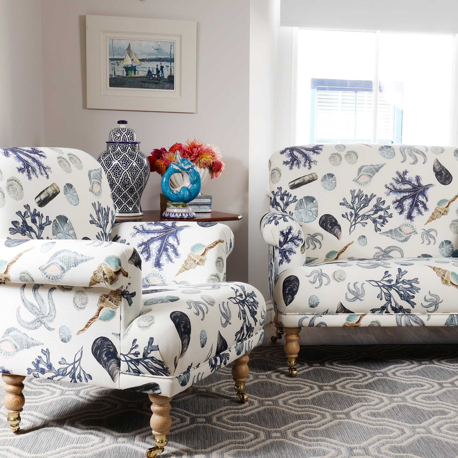 A bright and airy living room featuring a matching armchair and sofa upholstered in Cream Cornwall’s Rockpool fabric. The design showcases hand-drawn seashells, starfish, seaweed, and sand dollars in soft blues and neutrals on an off-white background. The space is styled with a patterned grey rug, a wooden side table holding books, a ceramic blue fish jug, a vase of fresh flowers, and a blue and white jar. Sunlight pours through a large window, enhancing the coastal, serene atmosphere.