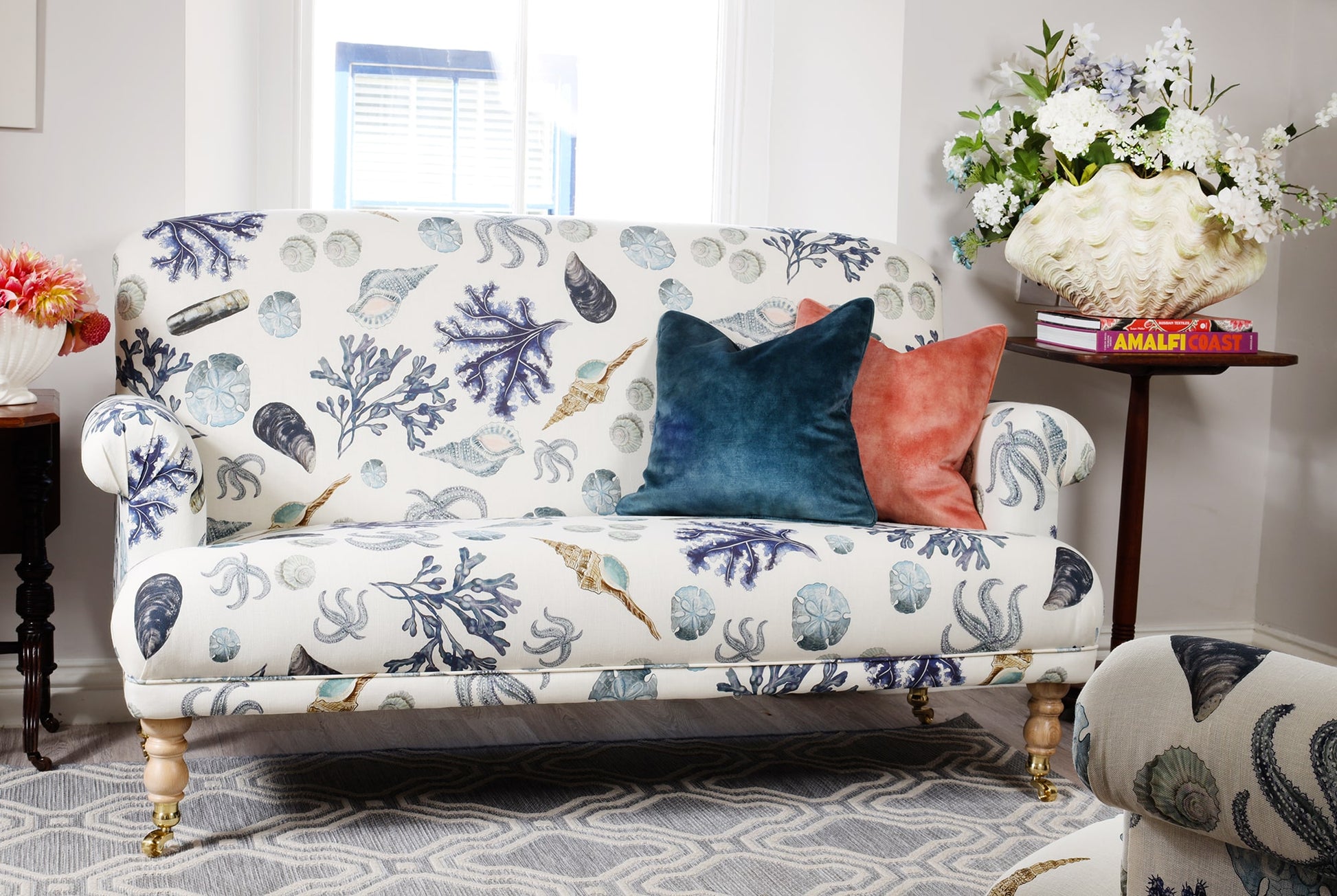 A bright coastal-inspired living room featuring a two-seater sofa upholstered in Cream Cornwall's Rockpool fabric in off-white, adorned with marine illustrations of seaweed, shells, and starfish in shades of blue and sand. The sofa is styled with two velvet scatter cushions—one in teal and the other in coral. A wooden side table holds books and a decorative clam shell vase overflowing with white and pastel flowers. Sunlight pours in from a nearby window, enhancing the tranquil, beachy atmosphere.