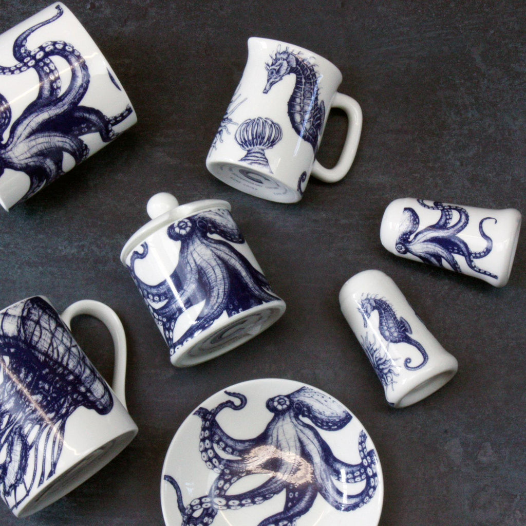 Bone china salt an pepper pots in our classic navy and white.Salt has seahorse, shell and an anenome on and the pepper has an octopus.They are both lying on a table also lying on the table is a seahorse jug and an octopus jam pot, octopus nibbles dish and some mugs all seen from an aerial view