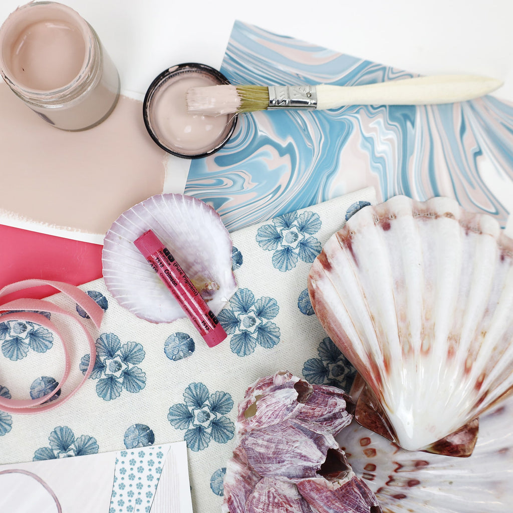 Flat lay of a coastal-themed mood board featuring a fabric with a blue sand dollar flower pattern, a marbled blue and blush wallpaper sample, seashells, a paintbrush dipped in blush pink paint, pink ribbon, and a bright pink crayon resting inside a scallop shell.