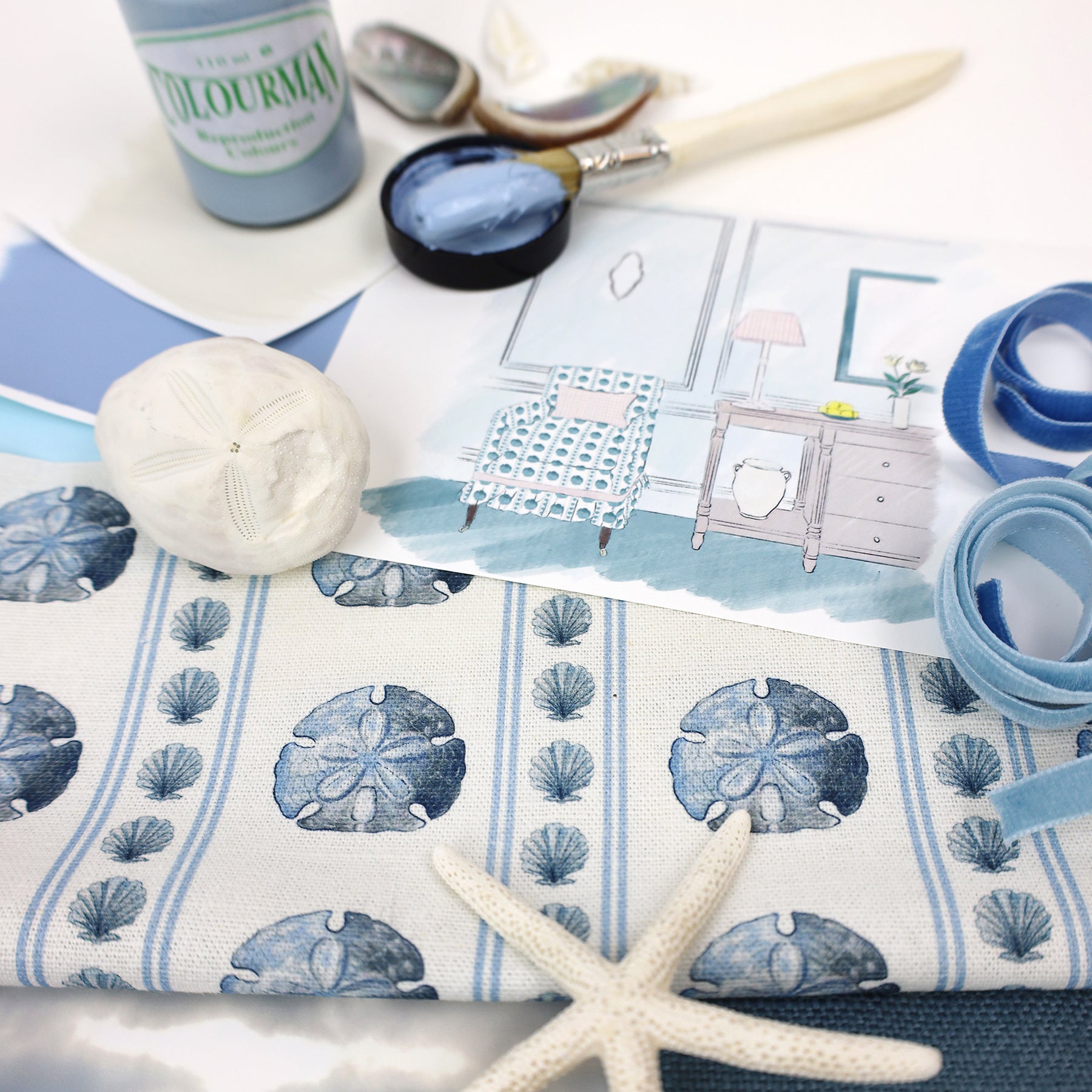 Coastal-themed mood board featuring a nautical fabric with blue sand dollars and seashells in vertical stripes. Surrounding elements include a watercolor interior illustration, blue velvet ribbons, a starfish, a white sea urchin shell, paint swatches, a brush dipped in light blue paint, and seashells. The scene captures a serene and creative beach-inspired design aesthetic.