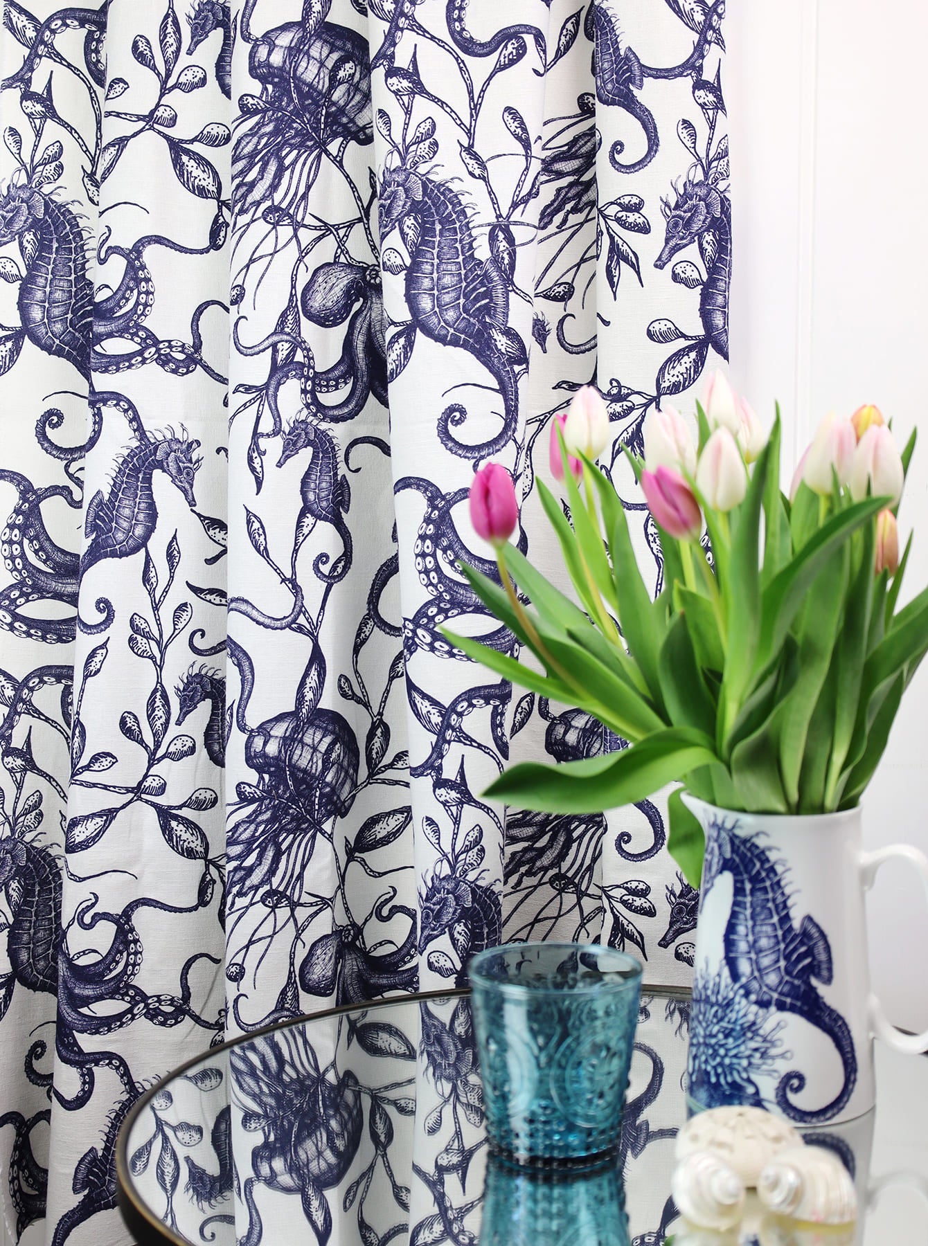 Close-up of a curtain made from Cream Cornwall’s Sea Creatures and Seaweed fabric in navy, featuring intricate illustrations of seahorses, octopuses, jellyfish, and seaweed. In the foreground, a glass-topped table holds a deep blue glass tumbler, two seashells, and a white jug with a seahorse motif filled with fresh pink and white tulips, creating a coastal-inspired interior setting.