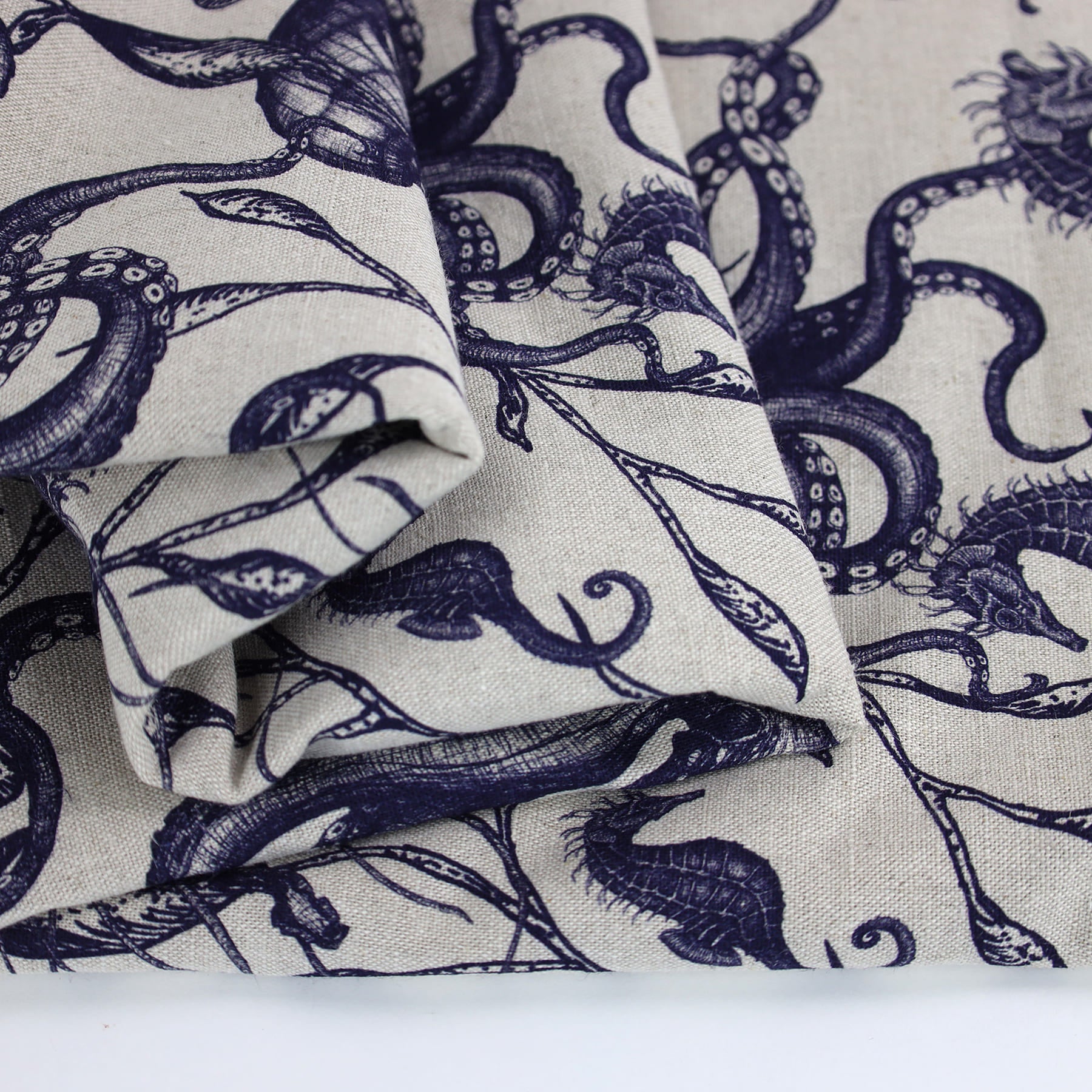 Close-up of folded Cream Cornwall fabric in natural linen, featuring detailed navy illustrations of sea creatures including seahorses and octopuses intertwined with seaweed. The intricate marine design evokes a classic coastal charm with a hand-drawn aesthetic.