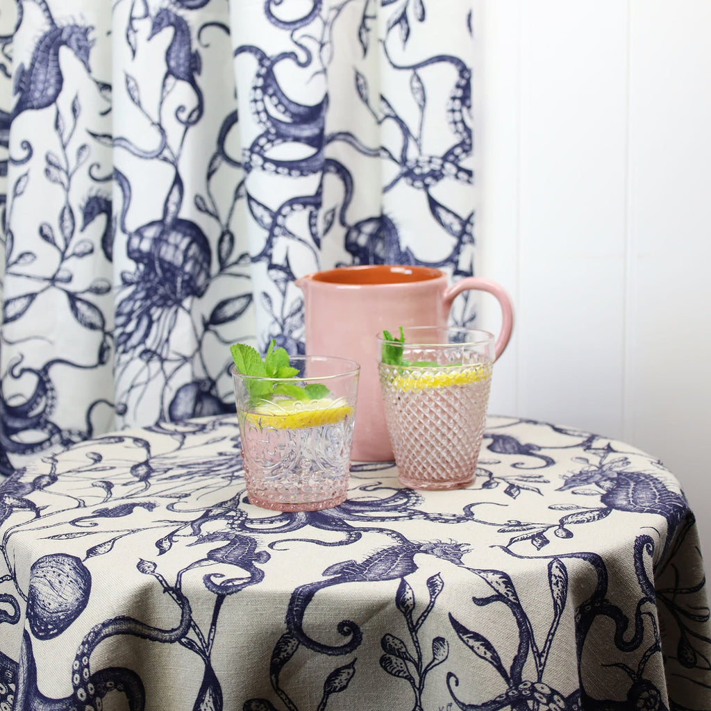 A charming table setting featuring Cream Cornwall’s Sea Creatures and Seaweed fabric in navy on a natural background, used as a tablecloth and curtain. On the table are two pink-tinted glasses filled with lemon water and mint, alongside a soft pink ceramic jug, set against a white wall for a fresh coastal look.