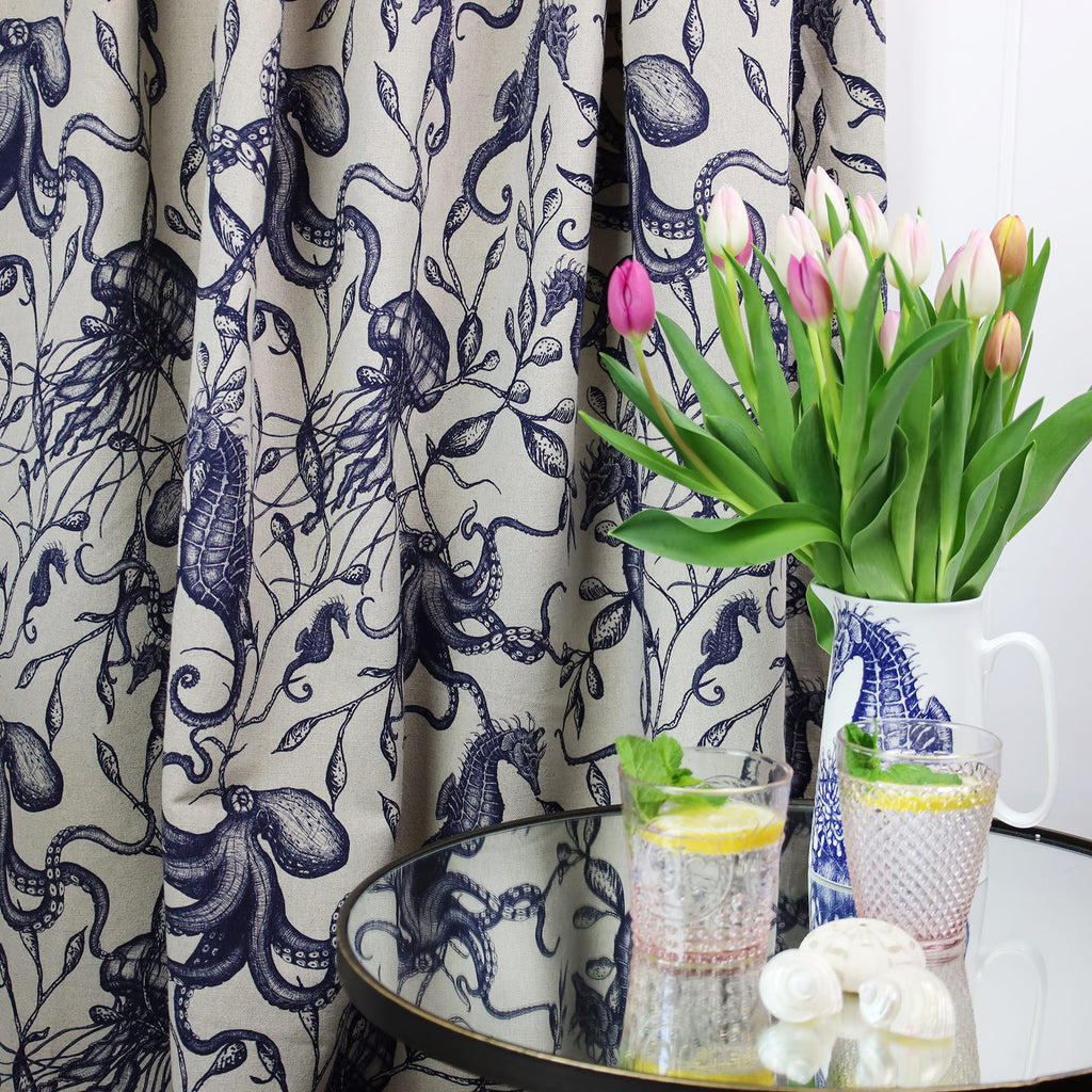 Nautical-themed interior scene featuring a curtain made of natural linen fabric with intricate navy blue illustrations of sea creatures and seaweed. In front is a glass-topped table holding two pink-rimmed glasses of lemon and mint water, decorative seashells, and a white jug with a seahorse design, used as a vase for pink and white tulips.