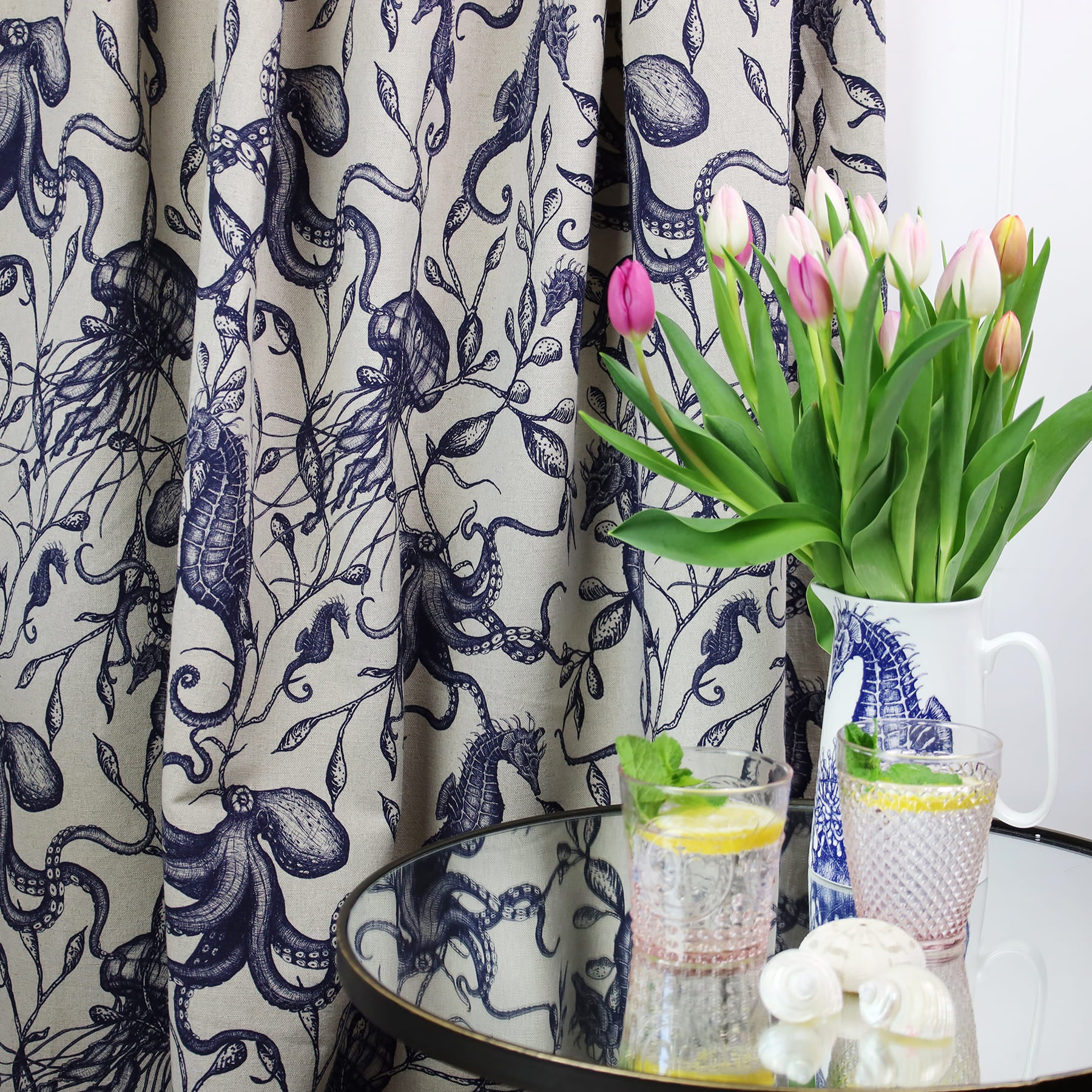 Nautical-themed interior scene featuring a curtain made of natural linen fabric with intricate navy blue illustrations of sea creatures and seaweed. In front is a glass-topped table holding two pink-rimmed glasses of lemon and mint water, decorative seashells, and a white jug with a seahorse design, used as a vase for pink and white tulips.