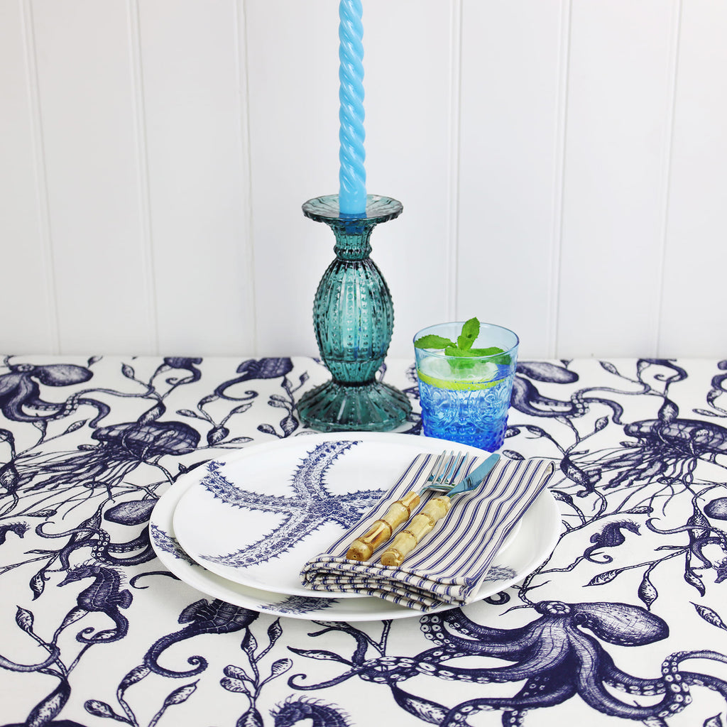 A coastal-themed table setting featuring Cream Cornwall’s Sea Creatures and Seaweed fabric in navy and white, used as a tablecloth. The scene includes a white plate with a blue starfish illustration, striped napkins with bamboo-handled cutlery, a blue glass of mint water, and a sea-green glass candlestick with a twisted blue candle, all set against a white panelled wall.