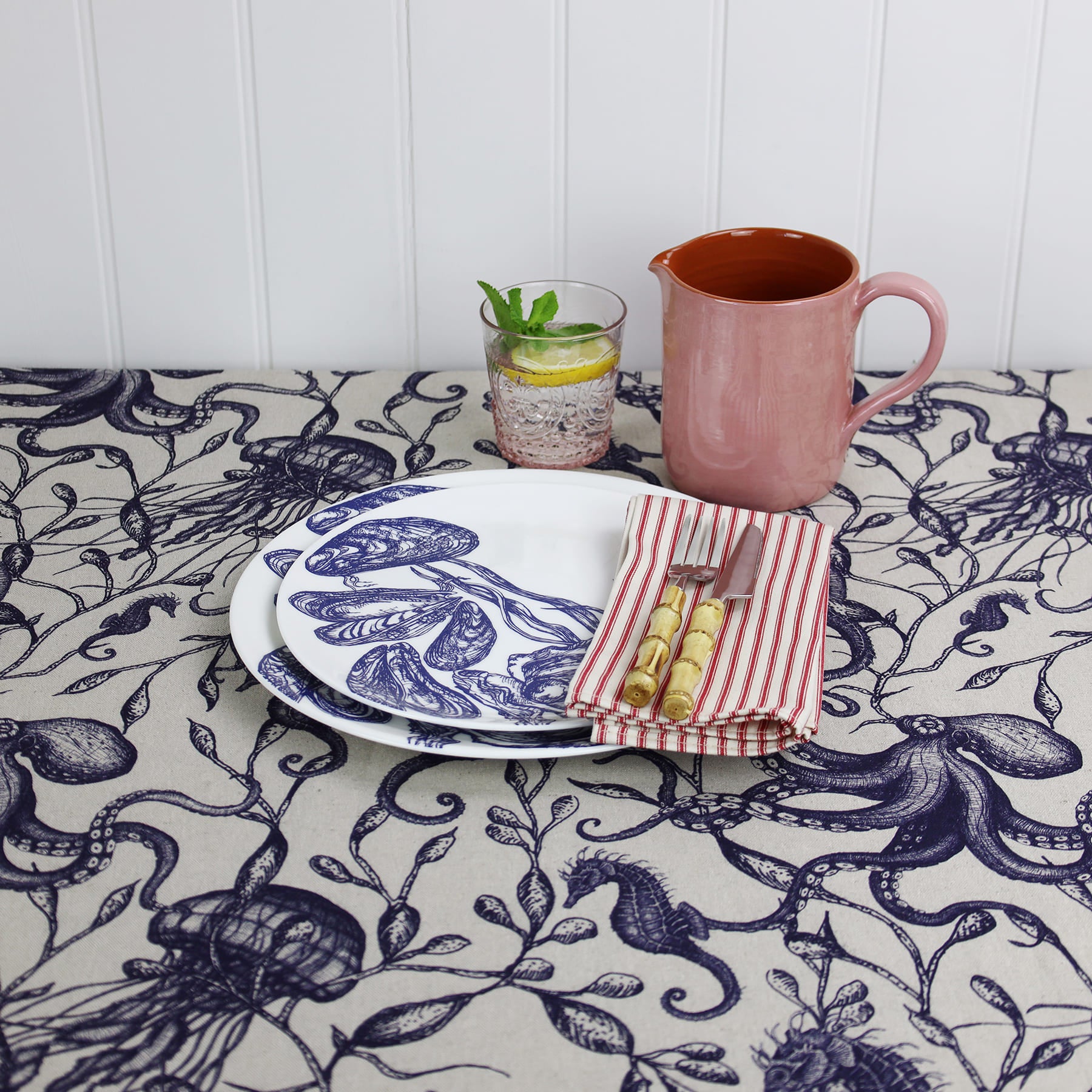 Coastal-themed table setting featuring a natural linen tablecloth adorned with navy blue illustrations of sea creatures and seaweed. The scene includes a white plate with a shell design, striped red and white napkin with bamboo cutlery, a pink ceramic jug, and a glass of lemon and mint water.