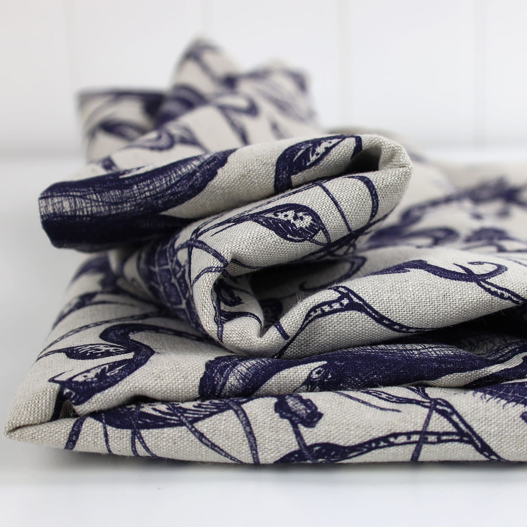 Neatly folded piece of Cream Cornwall fabric in a natural linen tone, featuring a hand-drawn navy blue marine design with detailed seaweed and ocean creatures. The fabric showcases a tactile weave and coastal elegance, ideal for upholstery or soft furnishings.