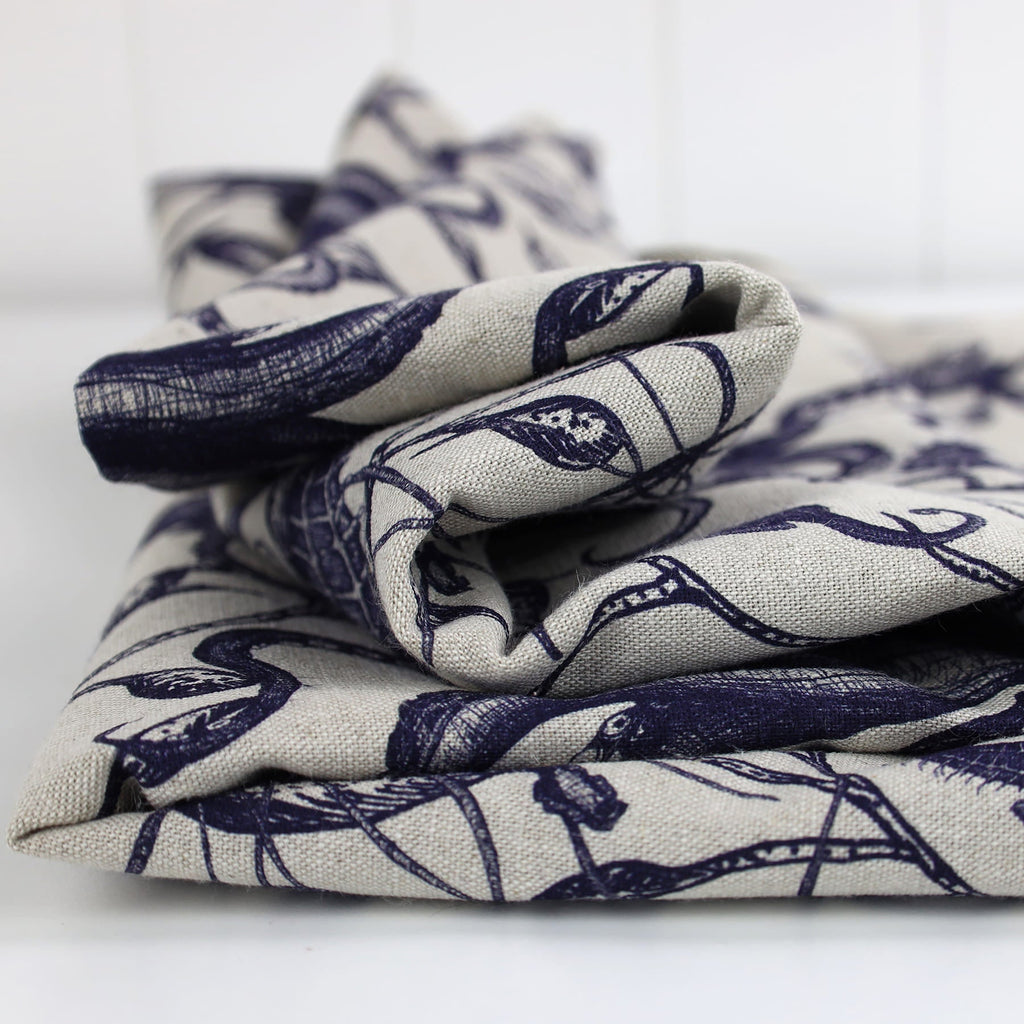 Neatly folded piece of Cream Cornwall fabric in a natural linen tone, featuring a hand-drawn navy blue marine design with detailed seaweed and ocean creatures. The fabric showcases a tactile weave and coastal elegance, ideal for upholstery or soft furnishings.