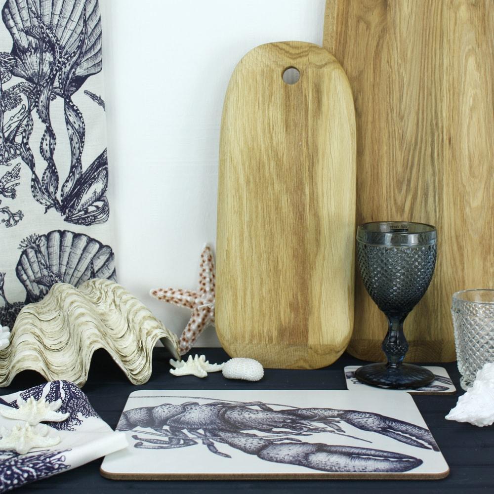 Lobster Design in Navy on a white Coaster with a matching Placemat.On the table are decorative shells and a matching napkin with wooden boards in the background